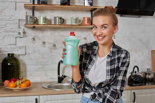Young smiling housewife on kitchen. Housekeeper woman holding bottle with cleaner liquid. Bottle copy space