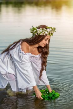 Beautiful black haired girl in white vintage dress and wreath of flowers standing in water of lake. Fairytale story. Warm art work. Sun flare