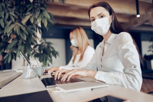 Two female colleagues working in office together wearing medical masks, coronavirus prevention