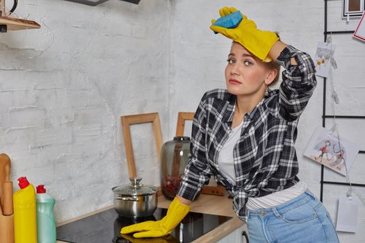 Blonde woman in protective gloves with rag cleaning electric stove at home kitchen. Girl washing black shiny surface of kitchen top. Concept of housework.