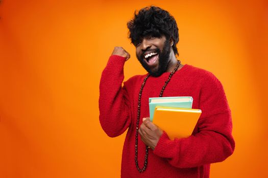 Successful african american male student holding books against orange background, close up