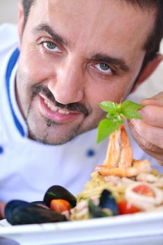 Handsome chef dressed in white uniform decorating pasta salad and seafood fish in modern kitchen