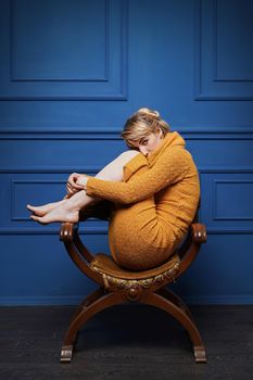 Studio portrait of a barefoot young woman in yellow pullover curling up in an old armchair on blue wall background.