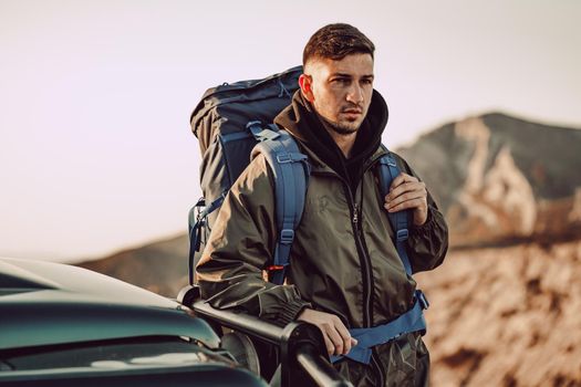 Portrait of a young traveler man in hiking equipment standing near his off-road car at dawn