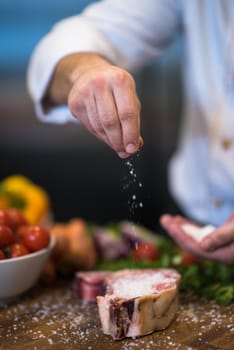 Master Chef hands putting salt on juicy slice of raw steak with vegetables around on a wooden table