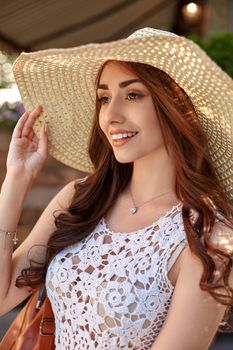 Sunny lifestyle fashion portrait of young pretty trendy girl posing at the city in Europe, summer street fashion, straw hat, laughing and smiling portrait.