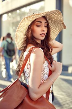 Fashionable brunette woman in a white dress, straw hat, walking in the street, posing near shop windows. Fashion summer photo