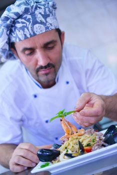 Handsome chef dressed in white uniform decorating pasta salad and seafood fish in modern kitchen