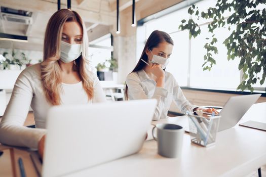 Two female colleagues working in office together wearing medical masks, coronavirus prevention