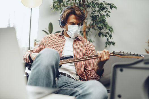 Young man plays guitar at home in medical mask. Coronavirus quarantine and self-isolation lifestyle concept