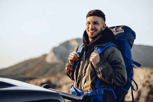 Portrait of a young traveler man in hiking equipment standing near his off-road car at dawn