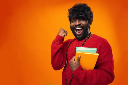 Successful african american male student holding books against orange background, close up