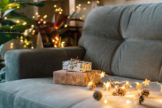 Christmas natural eco gift boxes with wooden label tag on gray couch background at cozy home. Glowing light bokeh, coniferous branch. Winter holidays. Zero waste, plastic free. Indoors plants