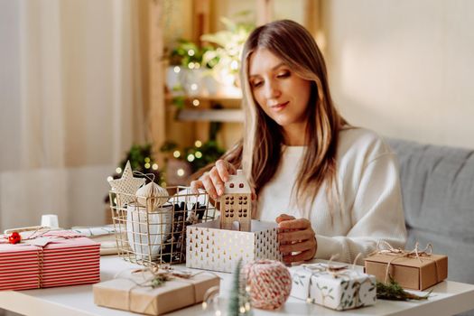 Woman wrapping Christmas eco natural gift boxes at home. Preparing presents on white table with decor elements, lights and items Christmas or New year DIY packing Concept.