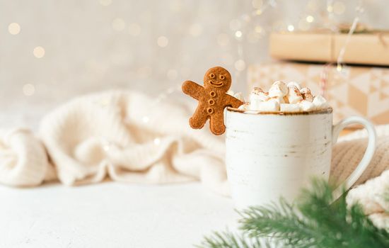Gingerbread man cookie in cup of hot cocoa or coffee with marshmallow, fir tree, gifts and warm cozy sweater. Christmas greeting card, lights background. Xmas holiday decorations with copy space.