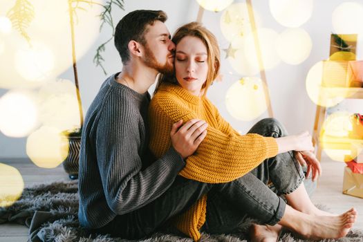 The guy gently kisses and hugs his girlfriend. Romantic couple of lovers against the background of a Christmas tree made of natural wood. Yellow bokeh.