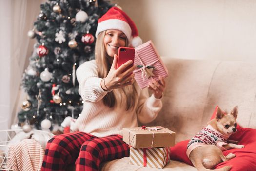 Young Woman and dog in sweater takes selfie portrait on the smartphone at winter holidays, enjoy Christmas time at home. Wrapping gifts and video chat at mobile phone.