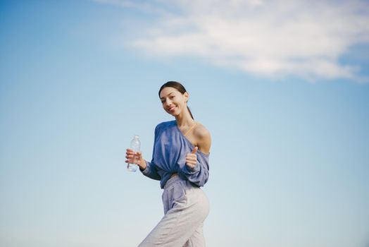 Beautiful girl training on a sky background. Woman in a blue sweater. Lady make a yoga. Girl with a bootle of water