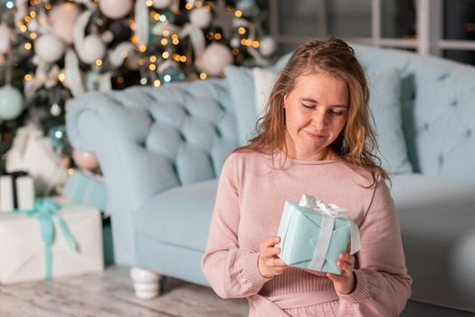 Beautiful middle-aged woman excited opening Christmas presents near the Christmas tree on Christmas eve