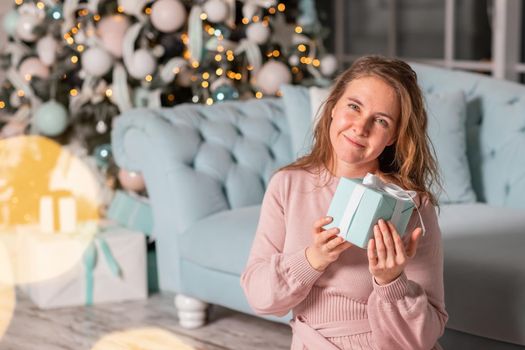 Beautiful middle-aged woman excited opening Christmas presents near the Christmas tree on Christmas eve