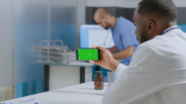 African american specialist doctor looking at mock up green screen chroma key smartphone with isolated display working at medical expertise in hospital office. Physician man analyzing sickness report