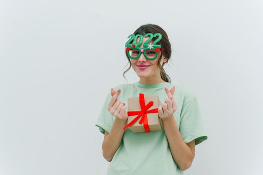 Modern young woman wearing 2022 glasses is holding a Christmas present and showing a Korean heart sign