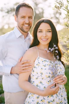 Man hugs pregnant woman holding wildflowers by the shoulders. High quality photo