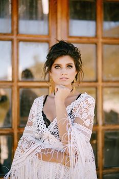 Young woman in a lace dress stands near a glass door, propping her head with her hand. Portrait. High quality photo