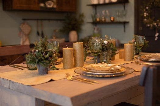 Close-up of beautifully decorated dining table with fir trees in vases and gold candles. Sets of plates for four people.