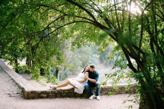 Man hugs woman while sitting on stone border in green park. High quality photo