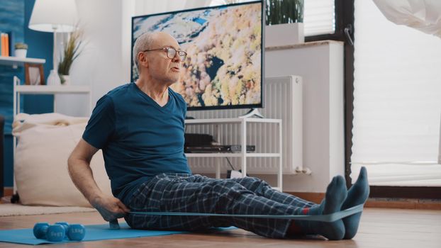 Aged man using resistance band to stretch arms and legs on yoga mat. Elder person pulling flexible elastic belt, stretching muscles and doing fitness exercise. Senior adult training