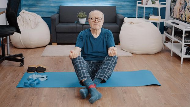 Aged man doing meditation while sitting in lotus position on mat. Senior person with eyes closed using yoga pose and concentration to meditate. Elder adult meditating for wellness.