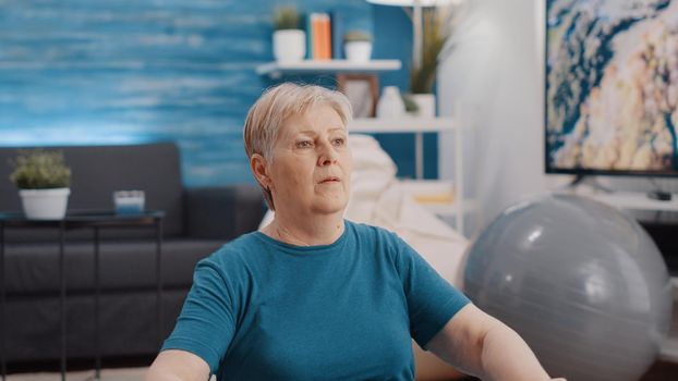 Handheld shot of elder person using resistance band to exercise and workout on yoga mat. Old woman pulling flexible elastic belt, training muscles with arm stretch for physical activity.