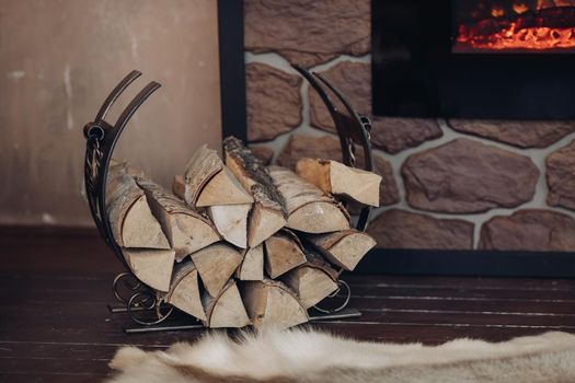 Decorative metallic holder with heap of wooden logs next to stony fireplace with burning logs.