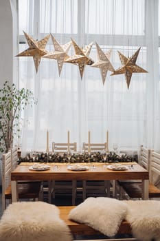 View over beautifully decorated dining table for six people with fir branch with pine cones in the middle of the table, served flutes and plates. Five decorative lamps in shape of stars over the table. Christmas dinner concept.
