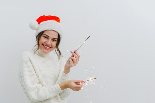 Young happy woman with sparklers celebrating and laughing over white background. Christmas and New Years celebration concept. Young girl wearing santa claus hat, copy space.