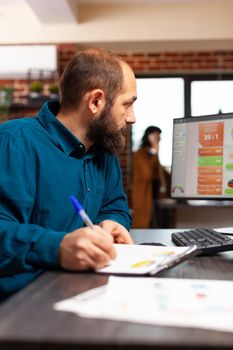 Businessman sitting at desk writing management strategy analyzing data charts using computer planning business presentation. Manager searching for solution brainstorming ideas in startup office