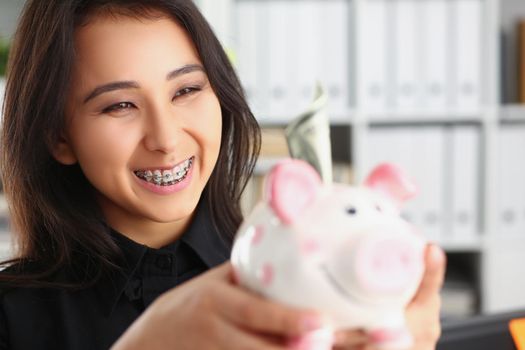 Portrait of pretty laughing young woman holding piggybank with banknote in it. Smiling brunette female save up for future. Investment, budget, fund concept