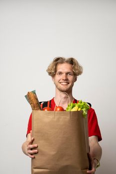 Portrait of pleased delivery man in red uniform smiling while carrying paper bag with food products isolated over white background