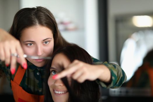 Portrait of concentrated professional hairdresser holding lock of hair and cutting with scissors. Happy female client sit in masters chair. Beauty concept