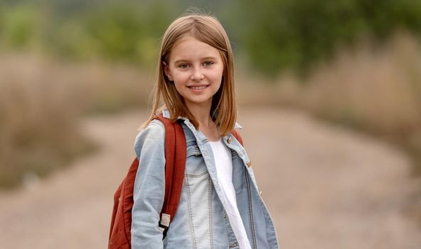 School girl with backpack looking at camera in the field with sunlight. Portrait of preteen child kid at nature