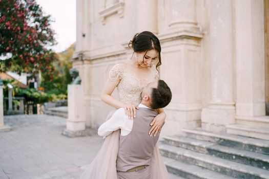 Groom holds bride in his arms in front of the marble facade of the old building. High quality photo