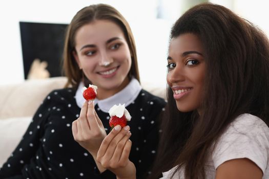 Portrait of smiling girls eating tasty fresh strawberry covered with whipped cream. Romantic date at home, best friends chilling. Lgbt, bff, lunch concept