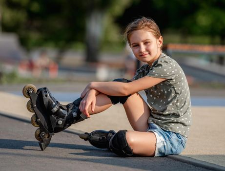 Beautiful preteen girl wearing roller skates sitting on ground and looking at camera. Portrait of pretty female kid skater
