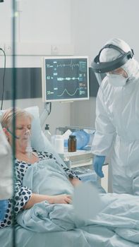 Medical staff in hazmat suits consulting aged patient with coronavirus. Sick woman in hospital ward quarantine receiving checkup visit from paramedics with protective uniforms and masks