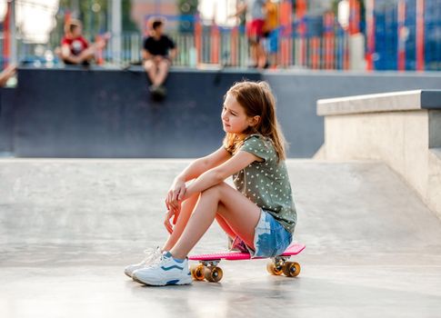 Preteen girl sitting on skateboard outdoors and looking back. Female skater child close to city riding ramp
