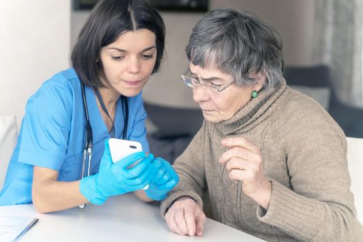 A nurse in a medical suit takes care and explains to an elderly patient how to use applications on a smartphone. Grandmother 80 years old, does not understand how to communicate with a doctor online.