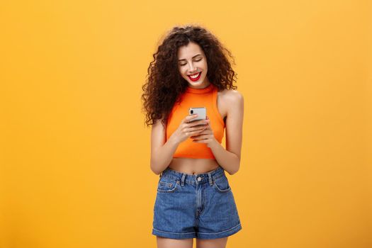 Girl spending time in internet texting friend messages. via smartphone laughing while looking at device screen standing happy and upbeat over orange background in cropped top and denim shorts.