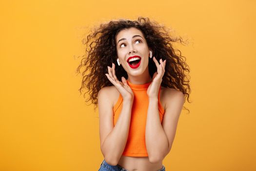 Waist-up shot of attractive silly curly-haired european female. in cropped top using wireless earphones touching earbuds and gazing at upper right corner delighted and carefree over orange wall.