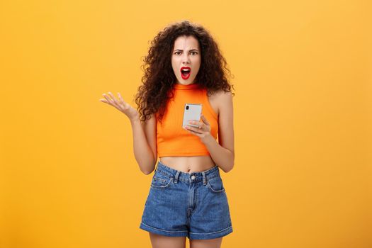 Portrait of displeased pissed and irritated caucasian female. with curly hairstyle in red lipstick and orange cropped top holding smartphone and gesturing with palm annoyed complaining on dumb message.